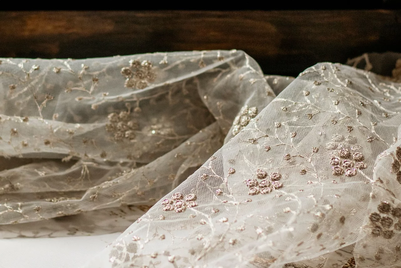 Close-up of a fine ivory tulle net in soft folds, raised floral embroidery and small reflective points worked across it.